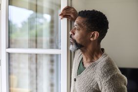 Older black man wearing a sweater is looking pensively out the window.