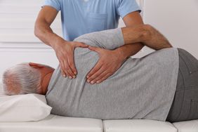 Older white man wearing a grey tshirt lying on his side receiving a back adjustment from a male chiropractor wearing blue scrubs.