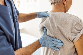 Older woman in a grey t-shirt is having her back examined by a healthcare provider wearing blue scrubs and gloves. 
