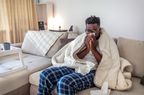 Black man sitting on couch wrapped in blanket blowing nose