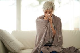 Woman with white hair wrapped in blanket blowing nose sitting on couch