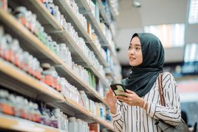Woman looks up information on phone while shopping in an aisle filled with supplements and vitamins.