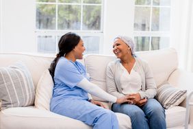 A healthcare provider and a woman undergoing cancer treatment laugh as they sit on a couch at home