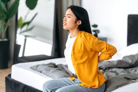 A woman sits on the edge of her bed holding her lower back in pain