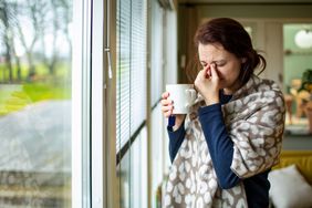 A woman stands at the window as she pinches her sinuses, eyes closed, with a mug in her hand and blanket wrapped around her