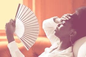 What Is Heat Intolerance? Symptoms, Risk Factors, and Ways to Relieve It , Tired Afro business woman suffering from heatstroke or hot summer flat without air-conditioner, touching her forehead, using waving fan, puts head on couch, sitting in living room at home. Overheating