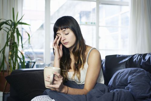 A woman sits with a mug of coffee in one hand, rubbing her closed eye with the other hand