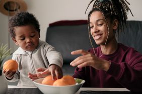 Young multiracial mother playing with oranges with her toddler son at home.