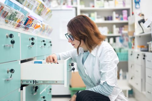 Woman in white robe and glasses looking through medications in drawer while working in modern pharmacy.