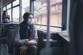 Worried young woman with protective face mask traveling with city transport and looking through the window.